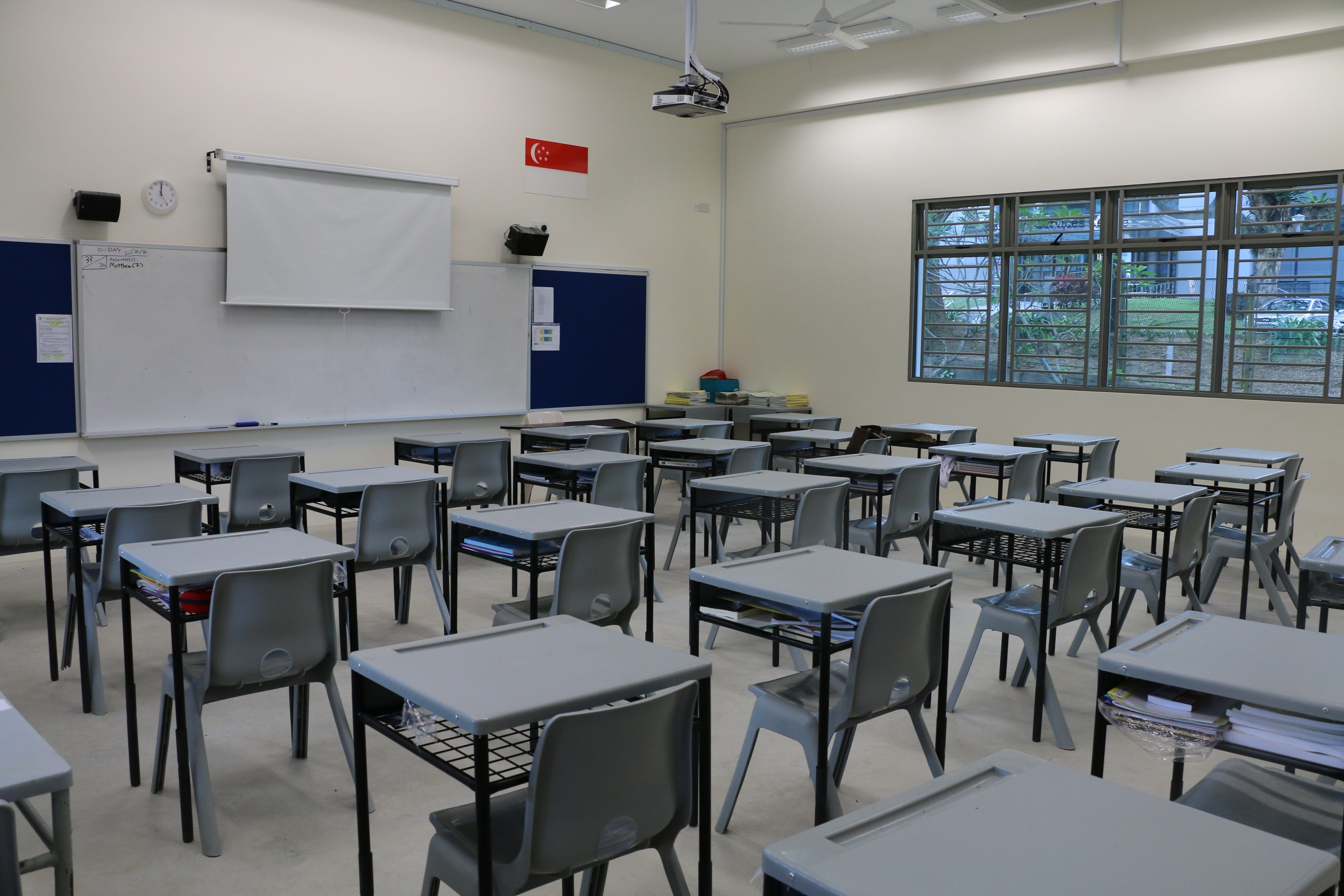 Empty classroom with rows of gray desks and chairs facing a white projection screen and blue bulletin board.